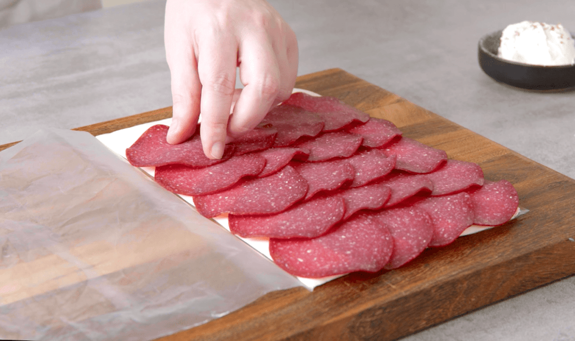 Salami Scheiben werden auf platt gerolltem Frischkäse verteilt